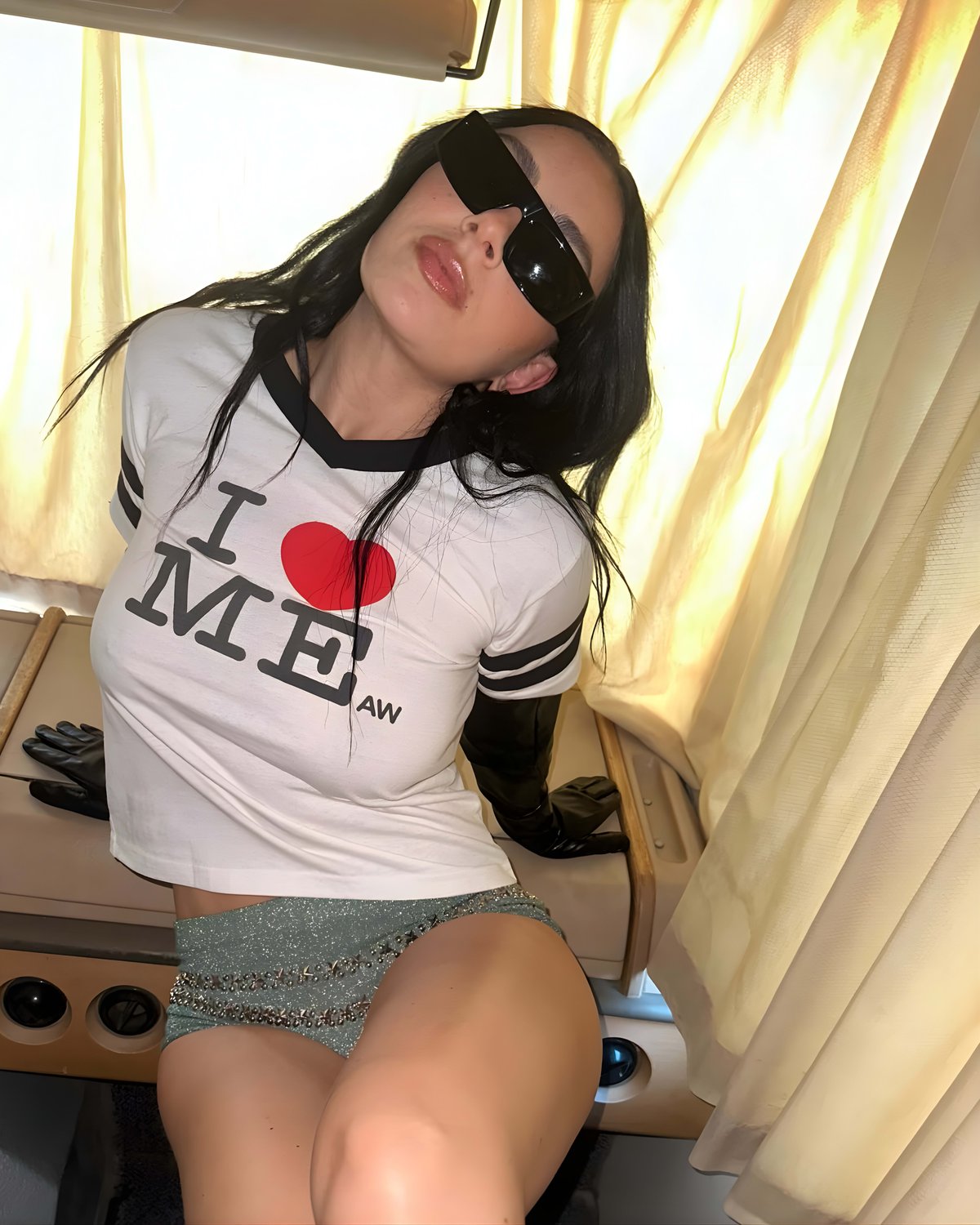 Charli XCX in black sunglasses, an "I love me" T-shirt and glittery shorts, leaning back by a curtained window