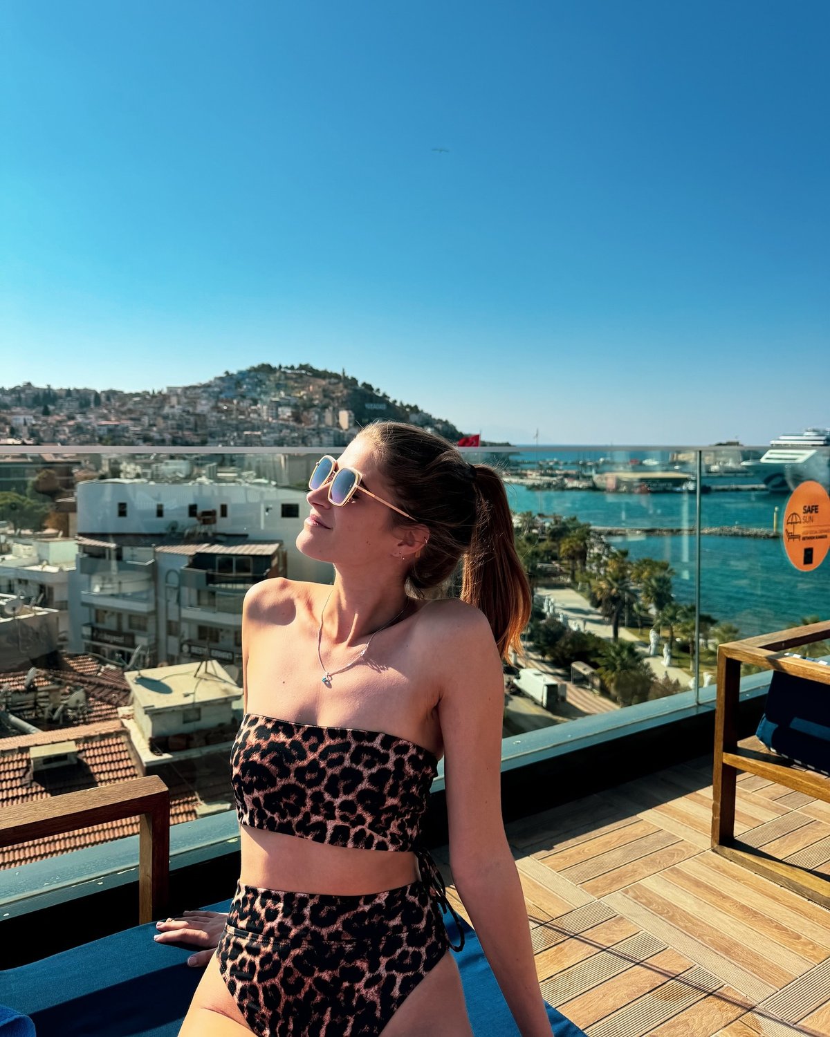 Sigrid Rijckoort on a rooftop terrace above a harbour town in a leopard-print bandeau bikini and sunglasses