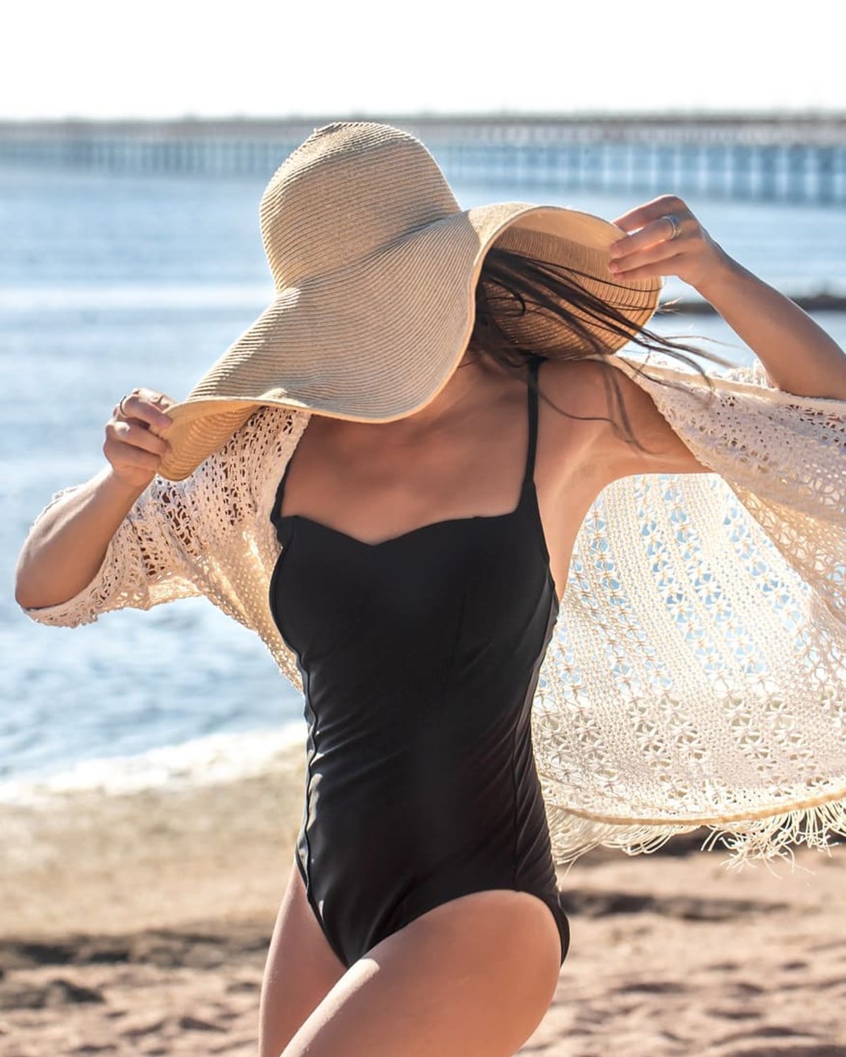 A woman wearing a black swimsuit, a wide-brimmed straw hat, and a light crocheted cover-up, with the ocean and a distant pier in the background