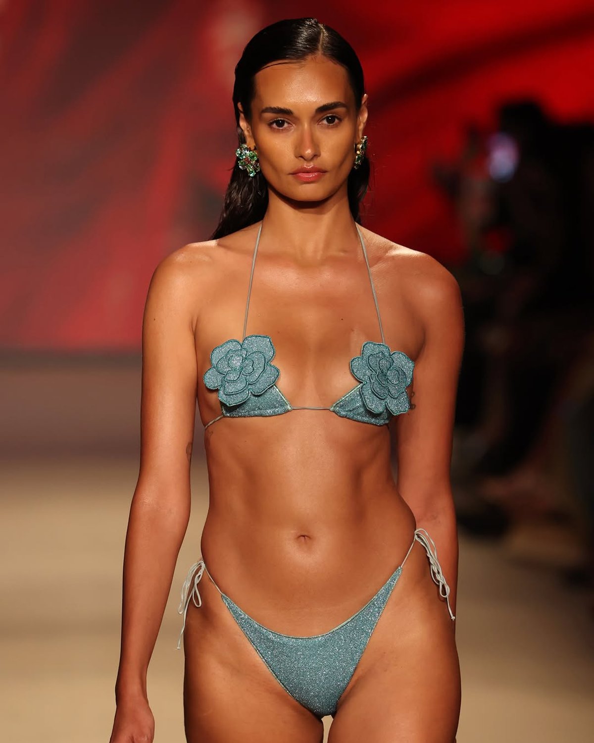 Model walking the runway in a shimmering teal bikini with floral appliqués, styled with bold earrings against a vibrant background.