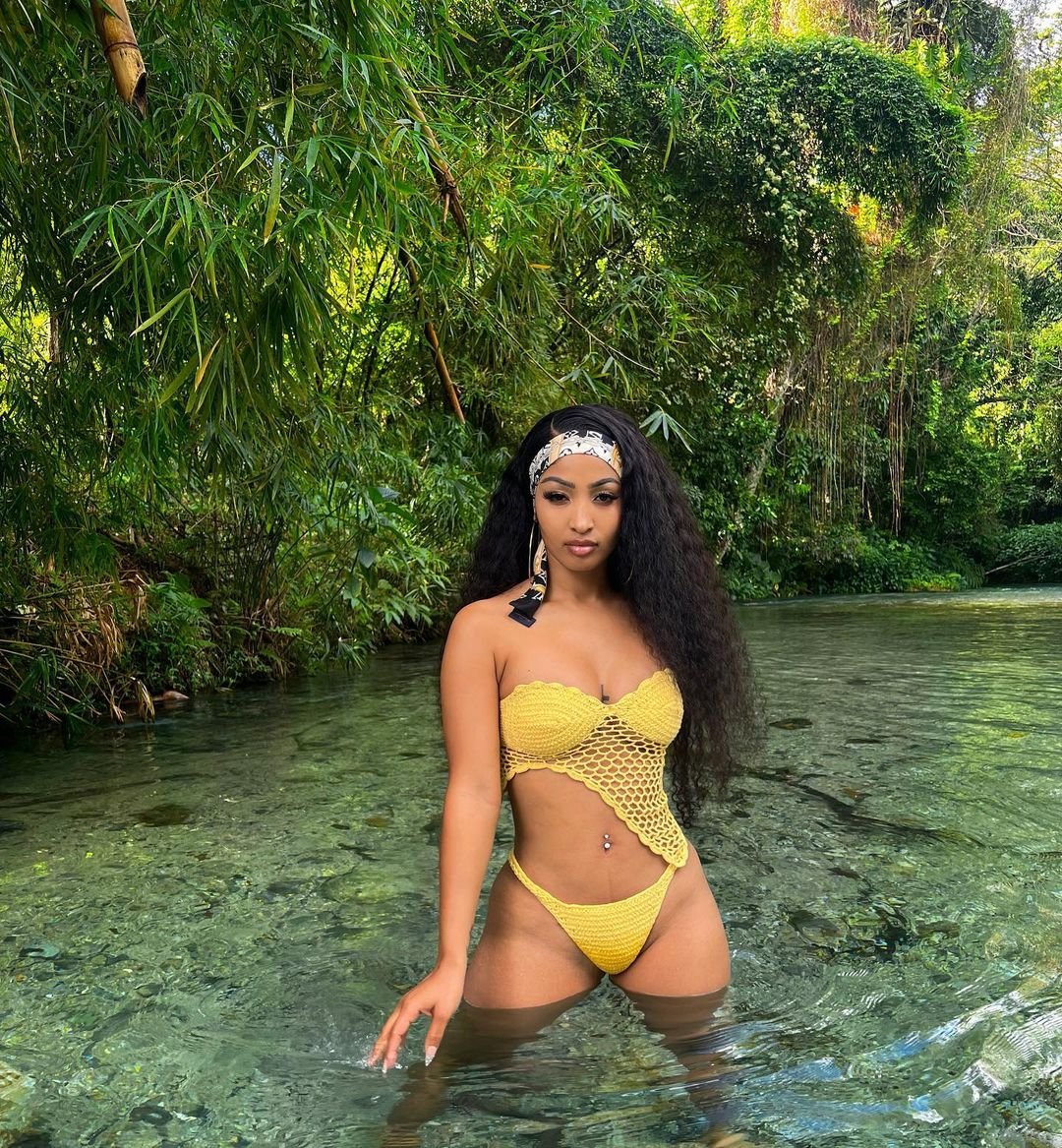 Shenseea in a yellow crochet bandeau top with a mesh panel and bikini bottoms, standing in a clear river under jungle trees