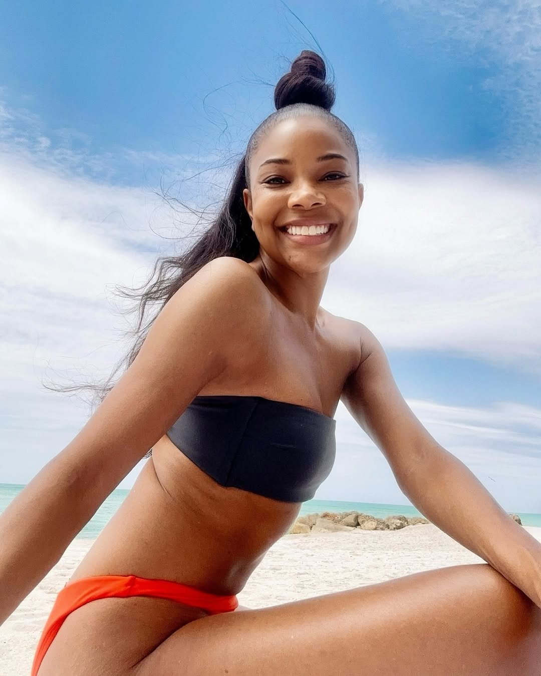 Gabrielle Union smiling on the beach in a black bandeau top and orange bikini bottoms