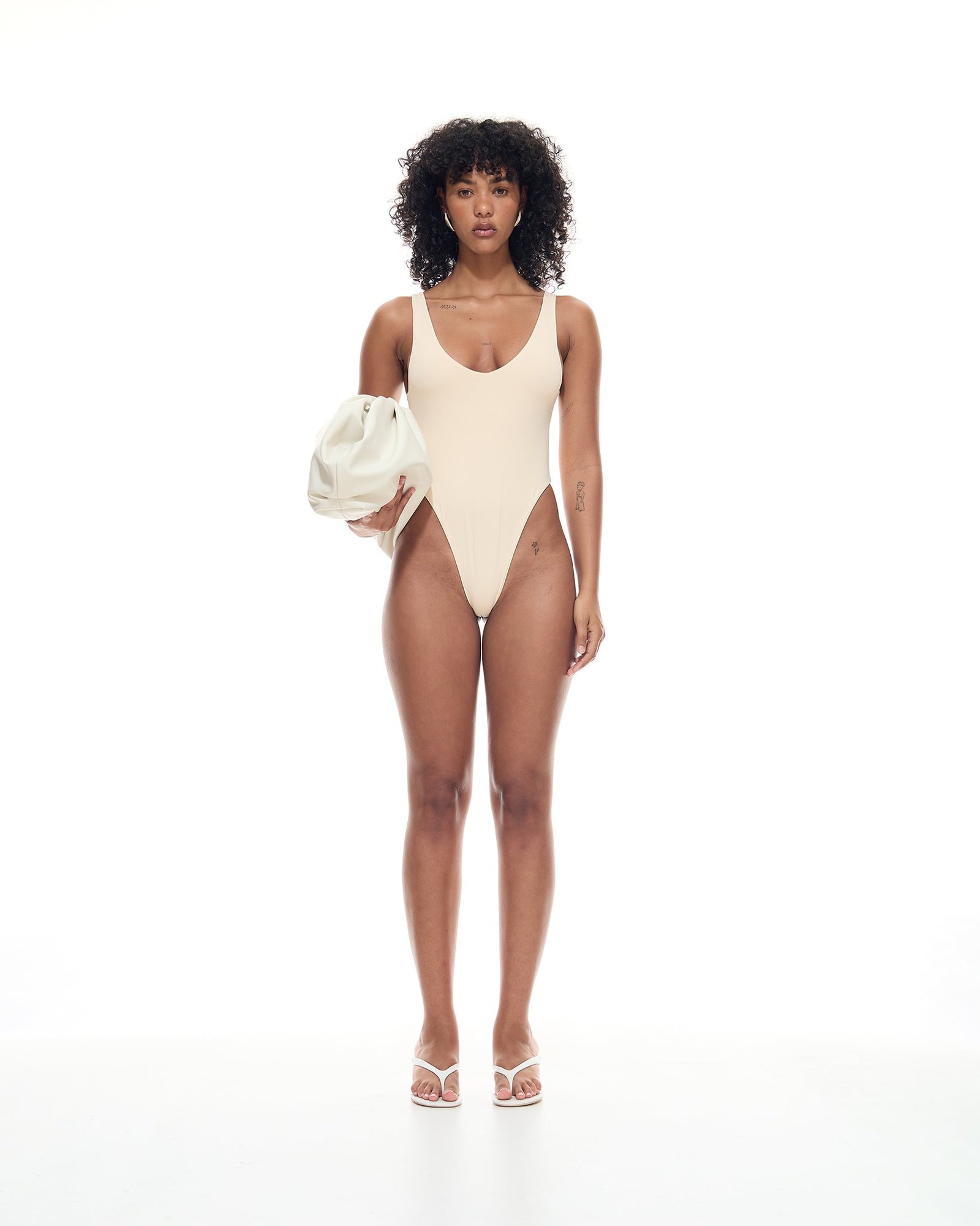 Model in the Amal One Piece in cream, a scoop-neck high-leg swimsuit, holding a white pouch bag