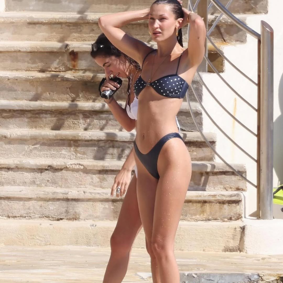 Model in a navy blue polka dot bikini, showcasing a toned figure, stands near stone steps by the water, adjusting her hair. A friend in a white bikini is in the background.