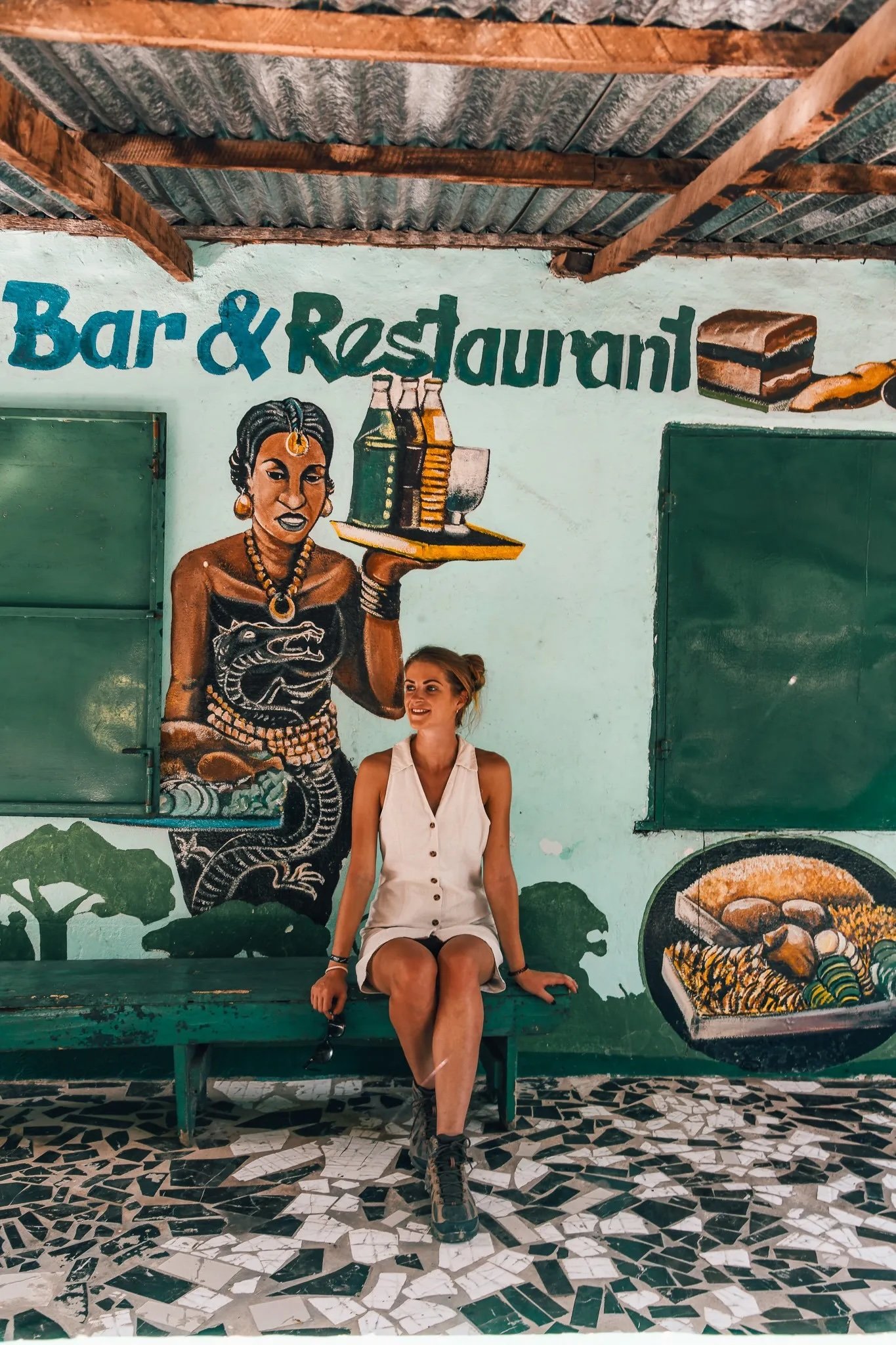 Sigrid Rijckoort in a white sleeveless romper, sitting on a green bench under a painted "Bar & Restaurant" mural
