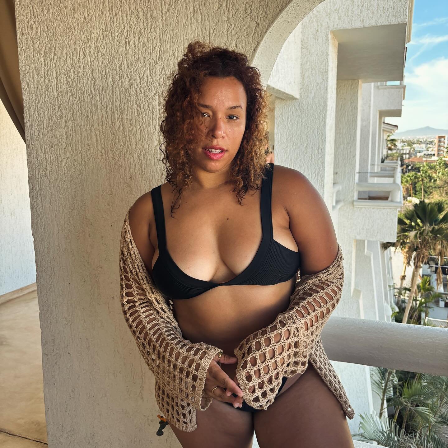 Woman on a hotel balcony in a black ribbed Tularosa bikini under an open tan crochet cover-up