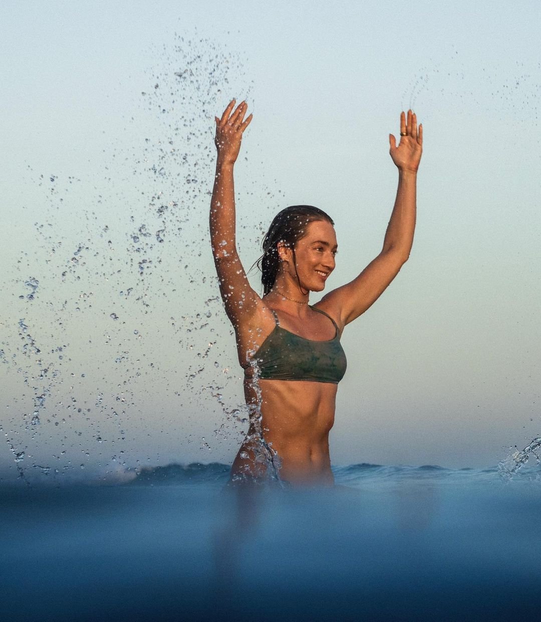 Sierra Quitiquit splashing water with her arms raised in a khaki bikini top, the sea at eye level