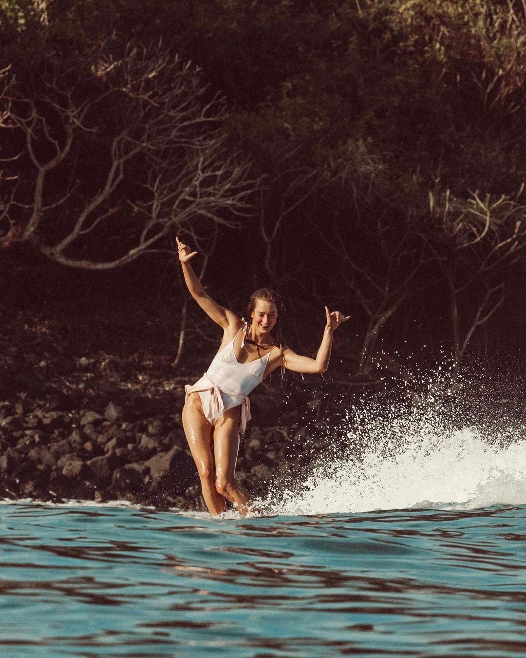 Sierra Quitiquit dancing on a surfboard on a wave in a white one-piece with a pink sarong tied at the hips