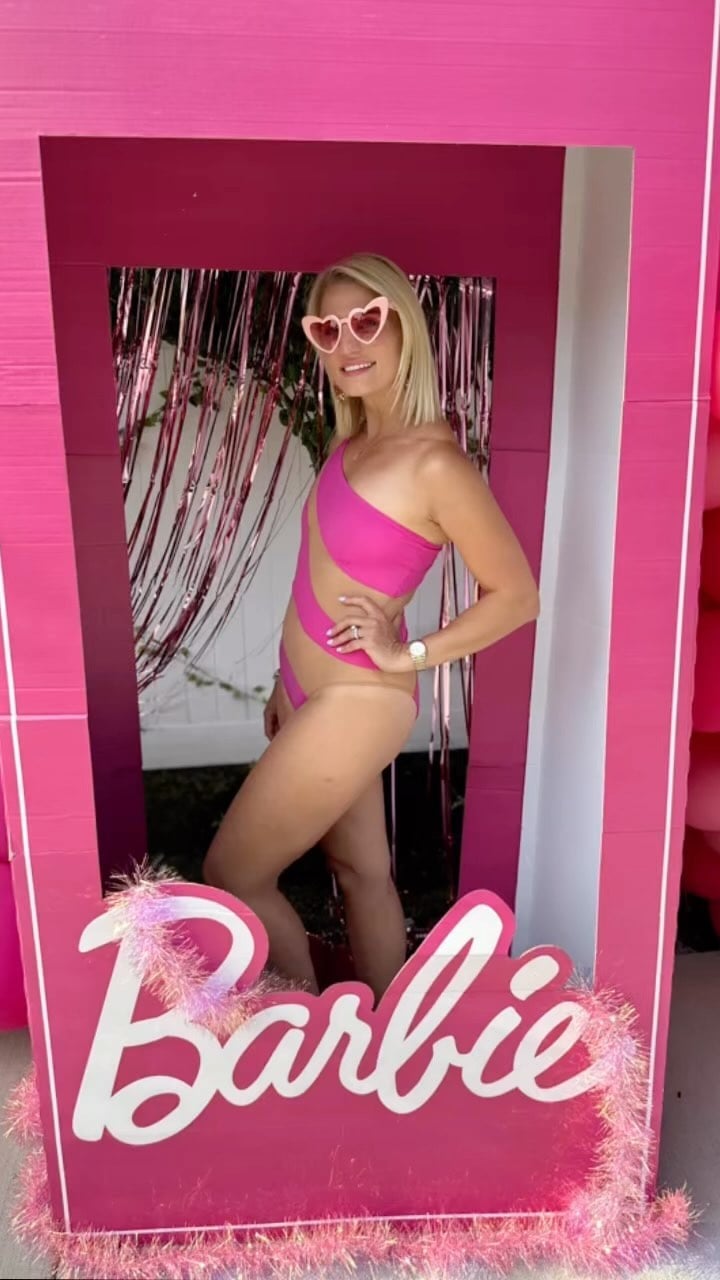 A woman in heart-shaped sunglasses posing inside a life-size Barbie box in a pink one-shoulder cut-out swimsuit