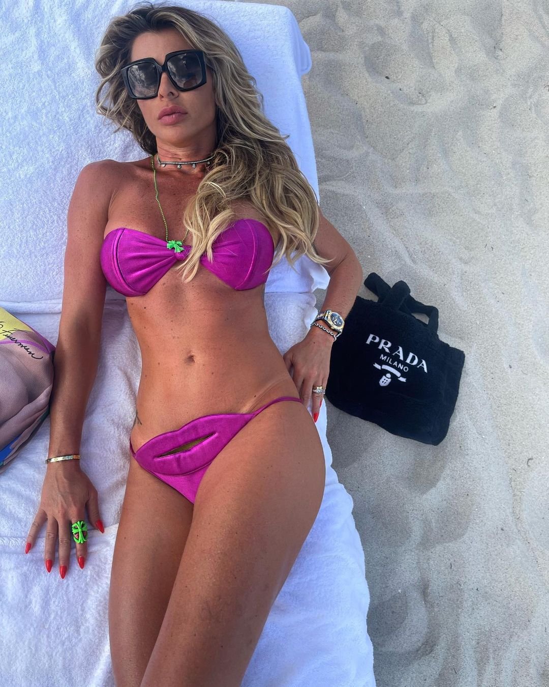 A woman in sunglasses lying on a white towel on the sand in a magenta strapless bikini with lip-shaped bottom