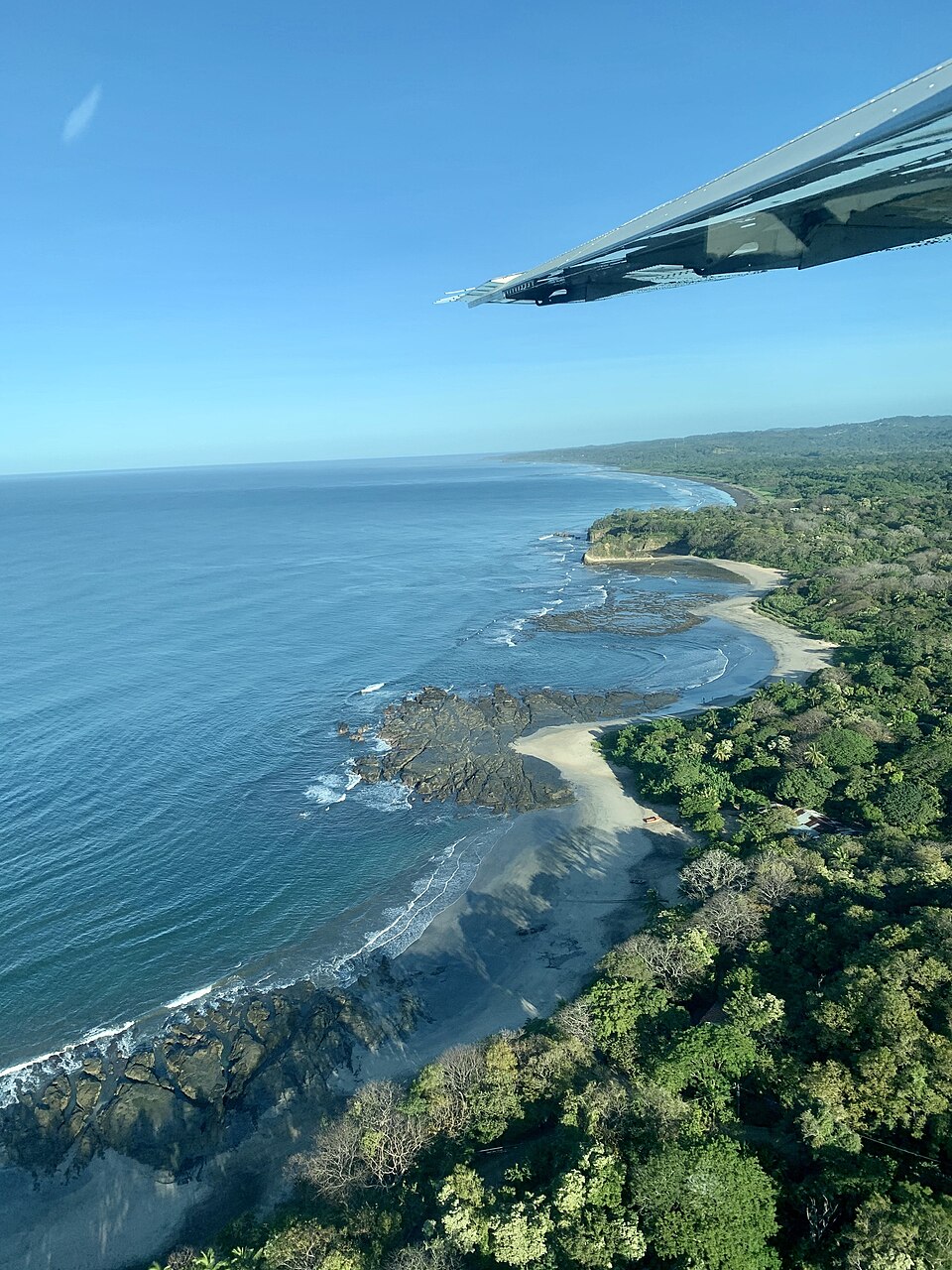 Aerial view of a serene coastline featuring rocky outcrops and soft sandy beaches, surrounded by lush green vegetation under a clear blue sky.