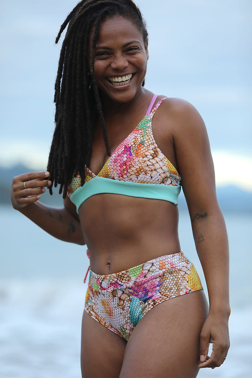 Smiling woman on a cloudy beach in the Magic Top and Pavones Bottom, a snakeskin-print bikini with a mint band