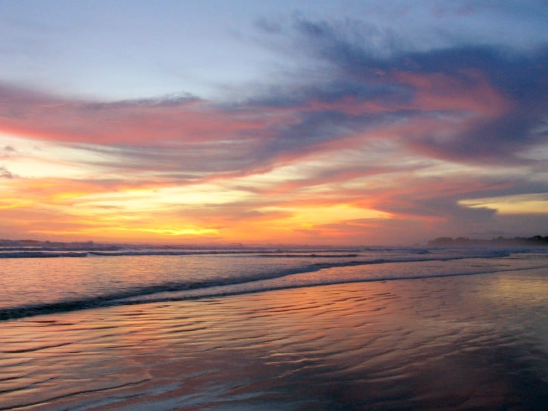 A stunning sunset over the ocean, with vibrant orange and purple hues reflecting off the wet sandy beach, creating a serene atmosphere.