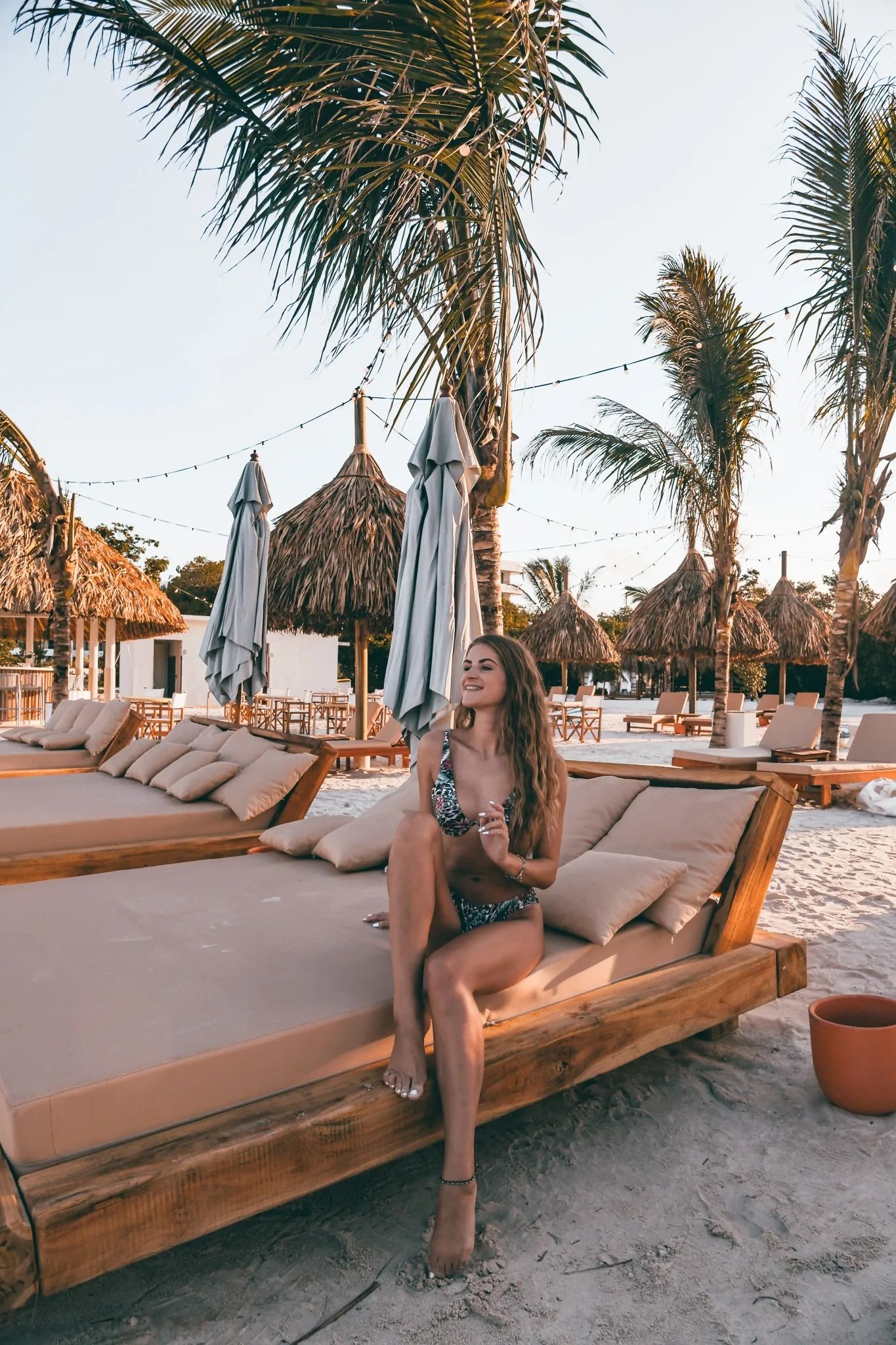 Sigrid Rijckoort sitting on a daybed at a beach club with thatched umbrellas and palm trees, in a black and white print bikini