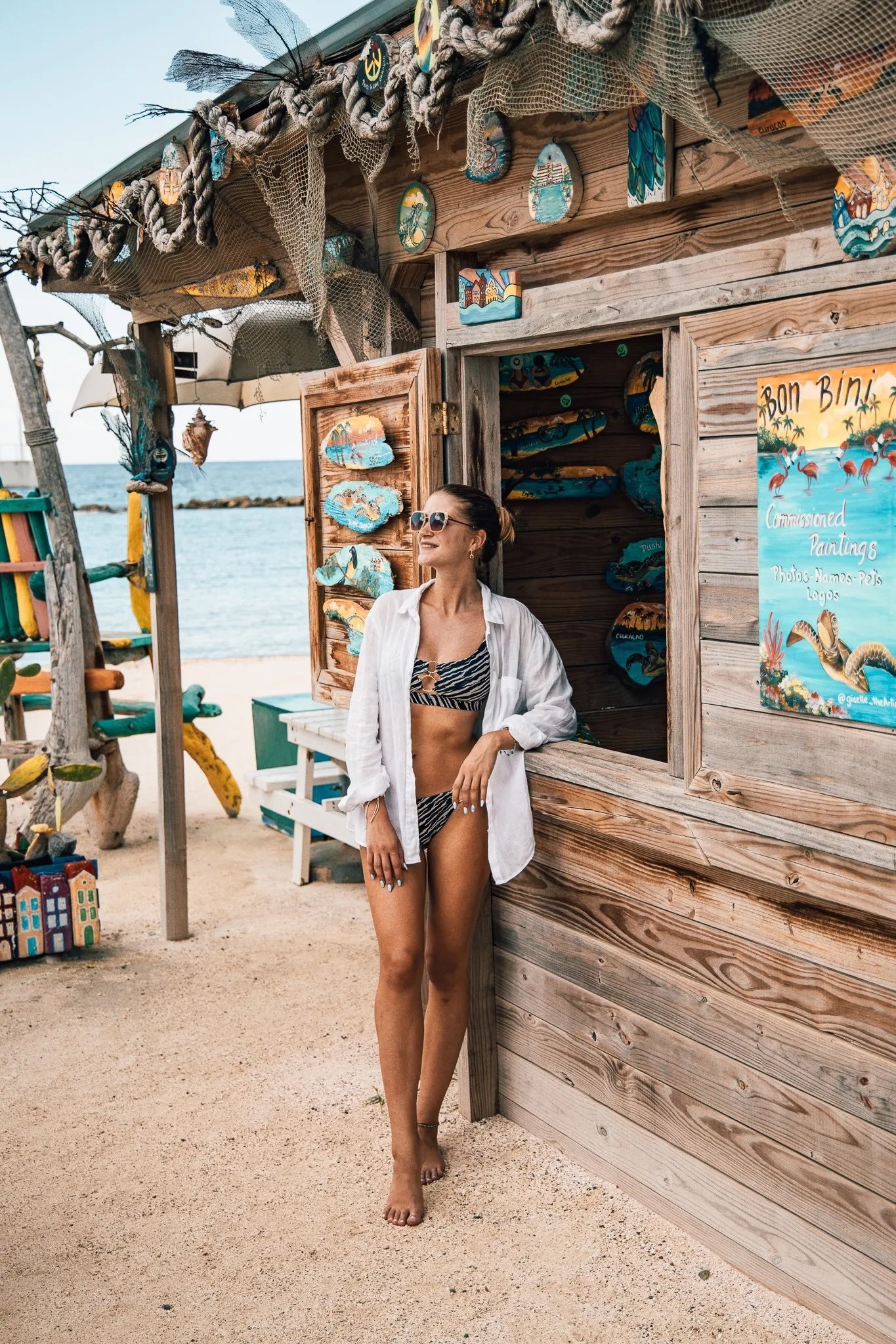 Sigrid Rijckoort in a zebra-print bikini and open white shirt, leaning on a painted wooden beach hut