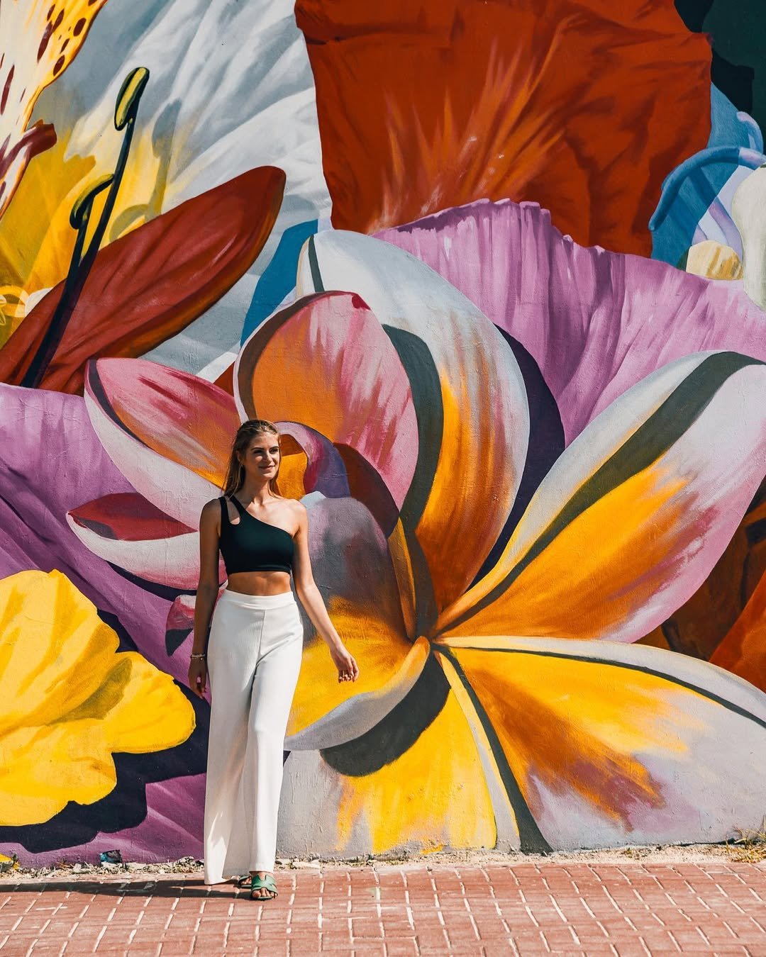 Sigrid Rijckoort in a black one-shoulder crop top and white wide-leg trousers in front of a giant painted flower mural