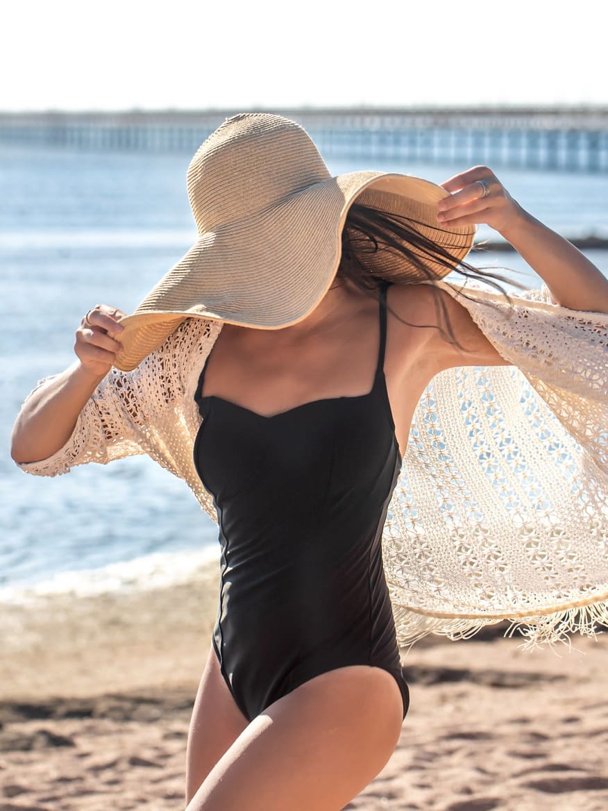 A woman wearing a black swimsuit, a wide-brimmed straw hat, and a light crocheted cover-up, with the ocean and a distant pier in the background