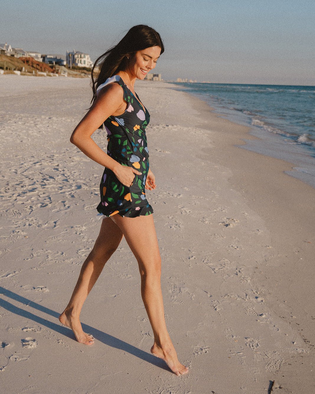 Woman walking barefoot along the shore in the Ginger Swim Romper, black with a pastel petal print and ruffled hem