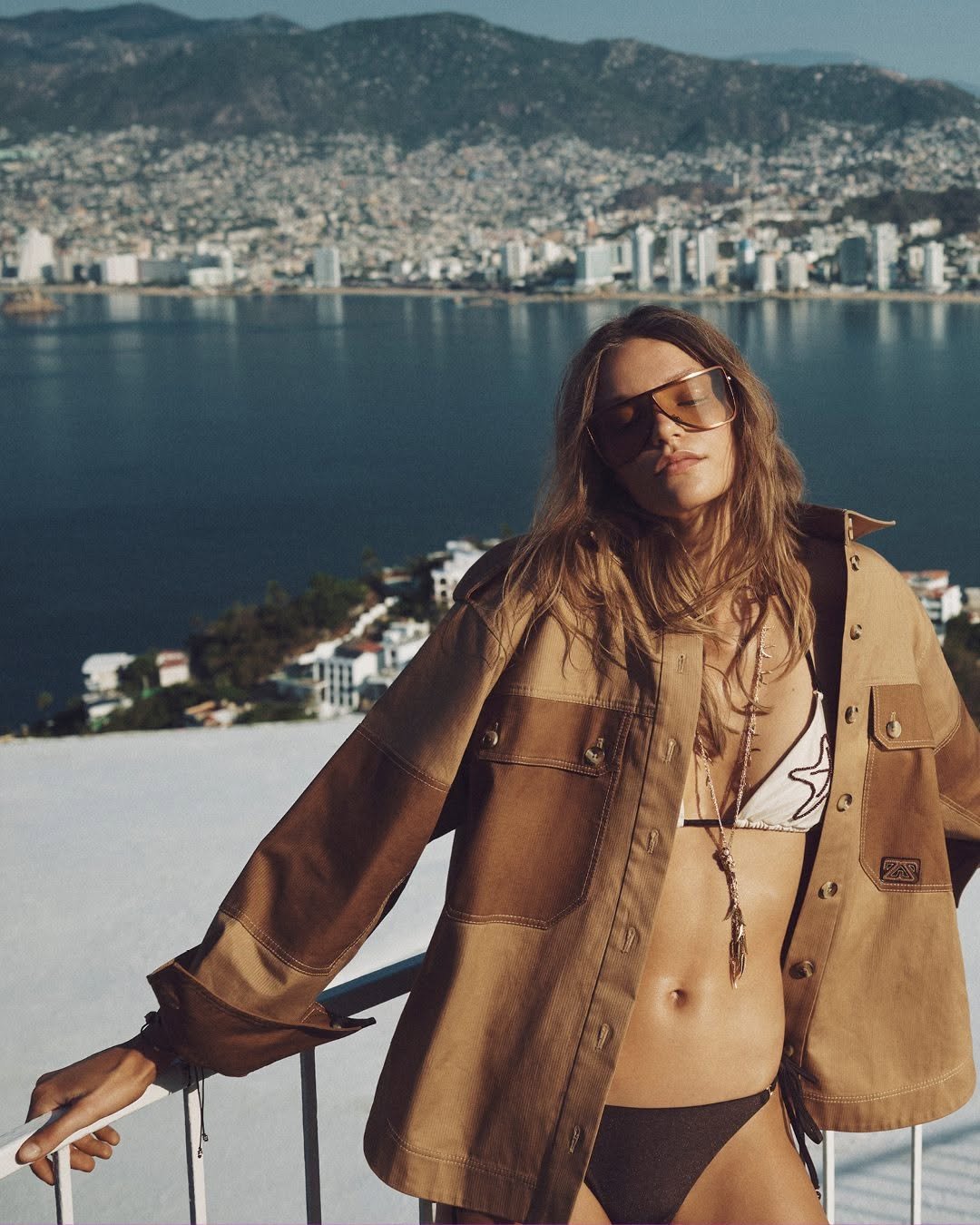 A model in a spliced bikini with beaded details poses on a balcony, wearing a brown oversized shirt and sunglasses, overlooking a seaside cityscape.
