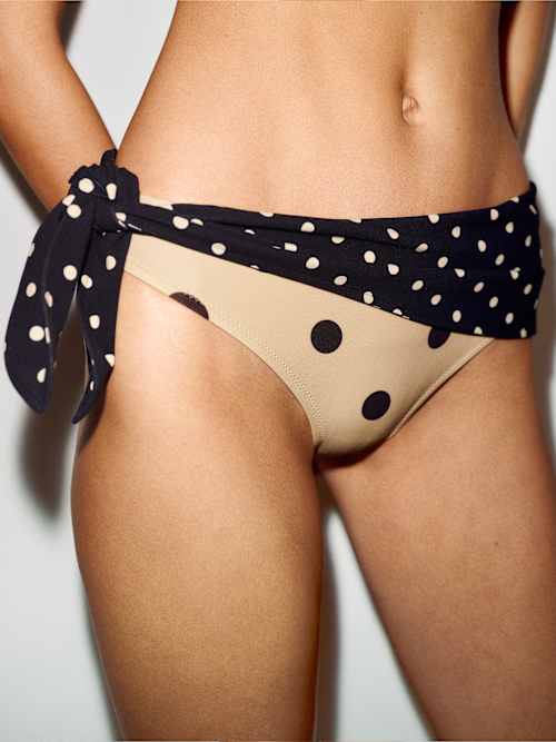 Close-up of the Riva Bikini Bottom in sand with big black dots, wrapped in a black polka-dot sash tied at the hip