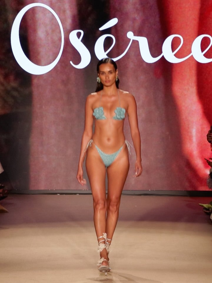 Model walks the runway in a turquoise Lumière Flower Kini featuring floral accents, paired with delicate ankle straps, against a vibrant backdrop.