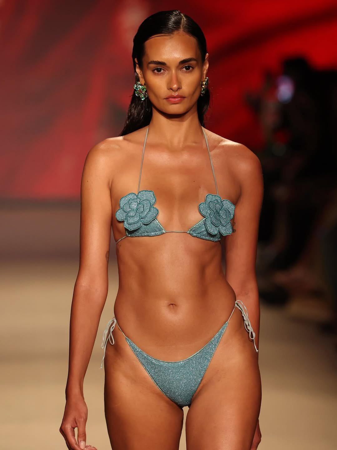Model walking the runway in a shimmering teal bikini with floral appliqués, styled with bold earrings against a vibrant background.