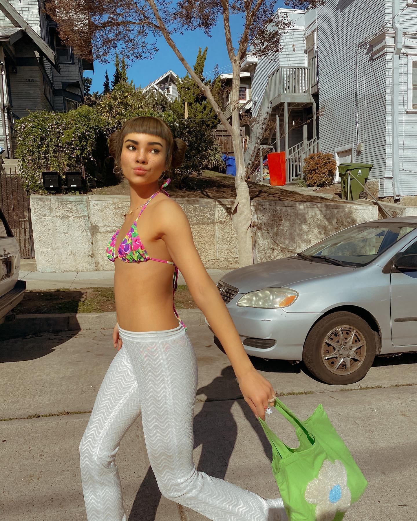 lilmiquela in colorful triangle bikini top and white zigzag pants, walking on a sunny residential street with a green bag.