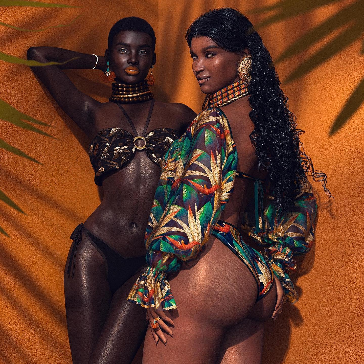 Two women in vibrant swimwear pose against an orange wall; one in a metallic bronze bikini with layered choker, the other in a colorful tropical cover-up and matching bikini, both with bold earrings and glossy, sun-kissed skin.