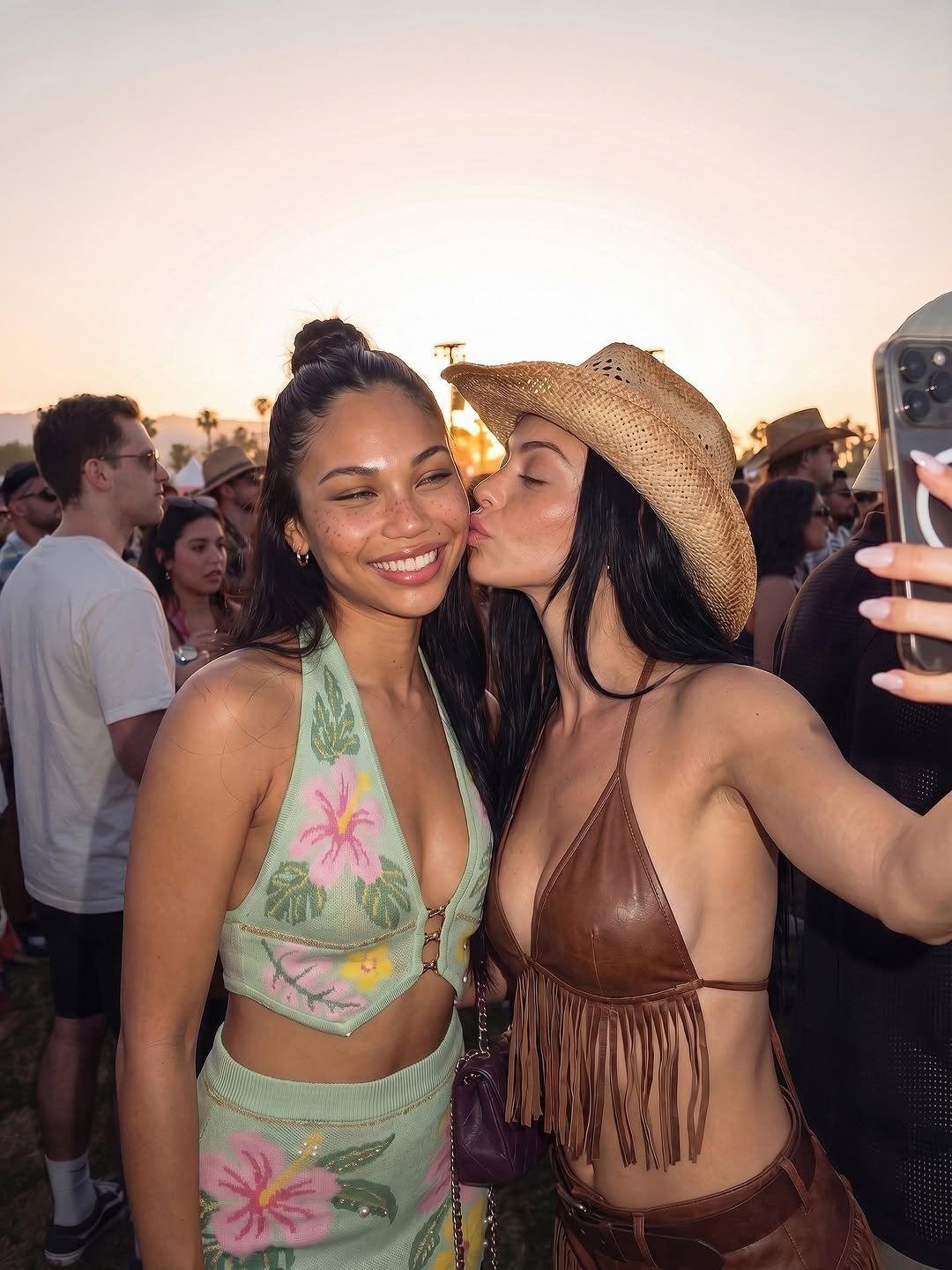 Two women pose for a selfie at Coachella, wearing vibrant festival outfits. One in a pastel floral halter top, the other in a tan fringed bikini, both smiling against a sunset backdrop.