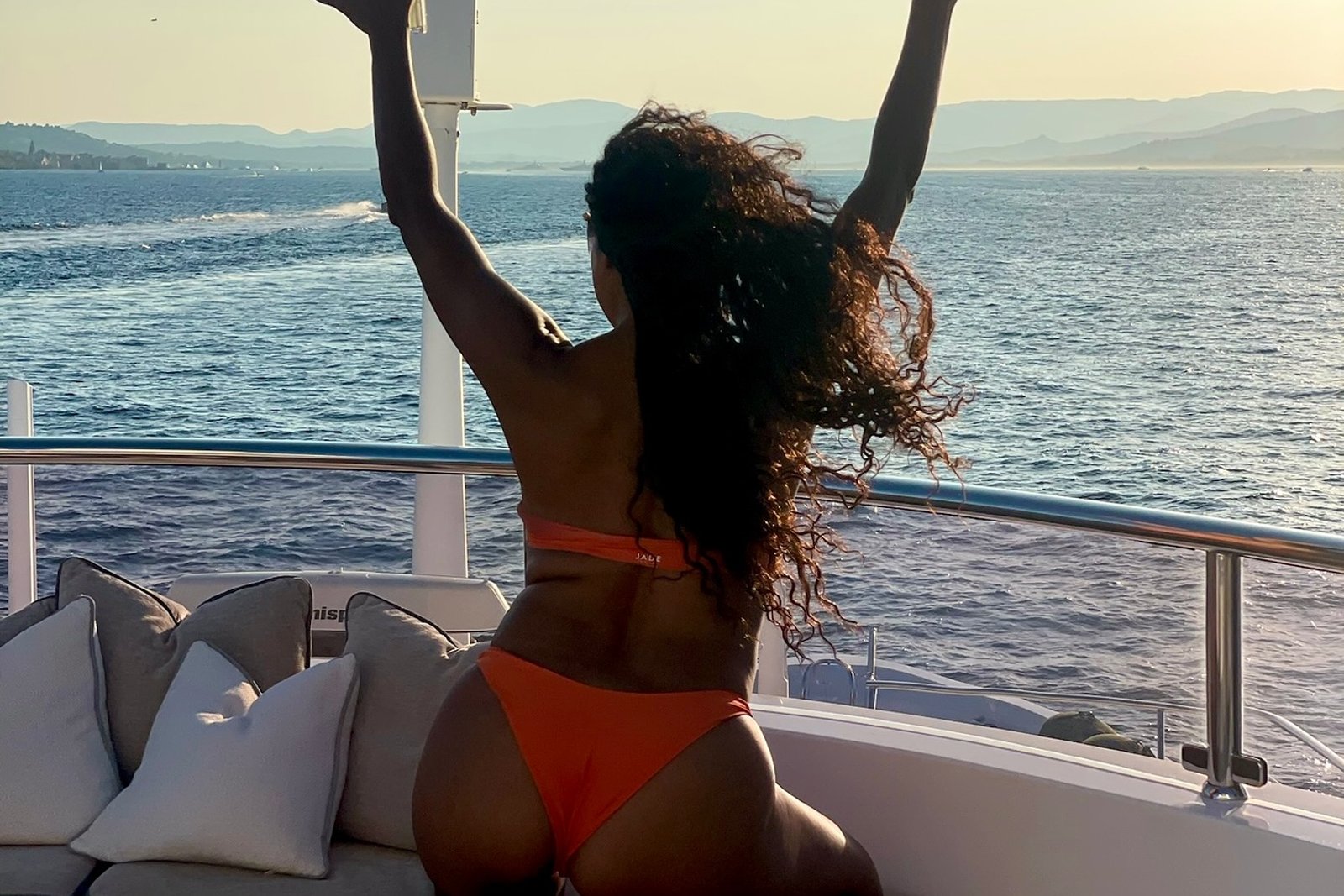 Gabrielle Union on a yacht deck at sunset in an orange bikini, seen from behind with her arms raised