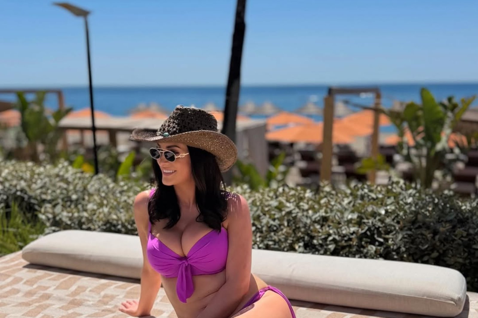 A woman relaxes in a vibrant purple bikini and straw hat, seated on a patterned lounge by the beach, with a sunny ocean backdrop.