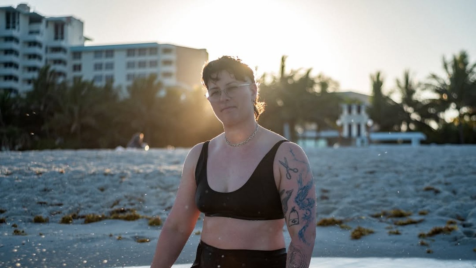 Woman in a black bikini top and swim shorts, kneeling in the surf at sunset