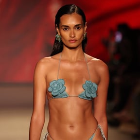 Model walking the runway in a shimmering teal bikini with floral appliqués, styled with bold earrings against a vibrant background.