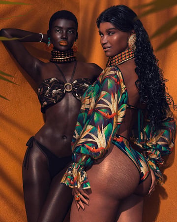 Two women in vibrant swimwear pose against an orange wall; one in a metallic bronze bikini with layered choker, the other in a colorful tropical cover-up and matching bikini, both with bold earrings and glossy, sun-kissed skin.